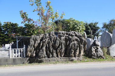 Asfalt yolun ortasında düz yüzeyi ve yuvarlak şekli olan büyük taş bir heykel var. Arkasındaki küçük bahçede kayaların üzerinde yetişen çeşitli ağaçlar var..