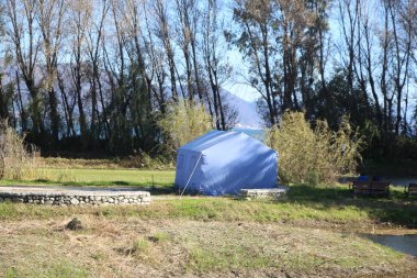 Mavi bir çadır çimlerin üzerinde yer alır, önünde ağaçlar ve göl manzarası vardır. Hemen yanımda boş bir park bankı var. Kış havasında, dışarıda güneş vardı..