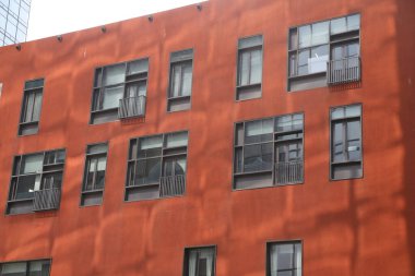 Siyah pencereli kırmızı boyalı bir bina, sokak seviyesinden çekilmiş bir fotoğraf. Bir tarafında turuncu bir duvar, diğer tarafında da pencereli bir bina var..