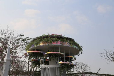 Beyaz metalden yapılmış, çatıda yeşil bitkiler ve gökkuşağı renkli çiçeklerle kaplı yuvarlak, çok katlı bir platform, yüksek irtifadaki bir endüstriyel park binasının önünde yer alır..