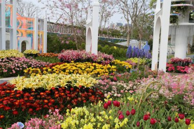 Bahar festivali çiçek ve bahçe gösterisi, parkta çeşitli renklerde çiçekler, büyük beyaz kemerler, bahar atmosferi, arkasında eğlence parkı olan renkli işaretler asılı.