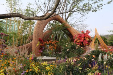 Çiçeklerle ve çeşitli renklerde bitkilerle dolu bir bahçe. Yeşil ağaçlar ve otlarla çevrili bambu şeritlerden yapılmış soyut bir ahşap heykel.