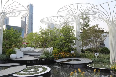 Çatı bahçesinde beyaz çelik çiçek stantları, yeşil bitkiler ve çiçekler, oturma alanının üzerindeki dairesel metal örtülü su çeşmeleri ve yemyeşil ağaçlarla çevrili..