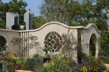 Kemerli penceresi olan modern bir bahçe duvarı çeşitli çiçek ve bitkilerle çevrilidir. Ortasına, müzik aletlerine benzeyen büyük bir heykel yerleştirilir.