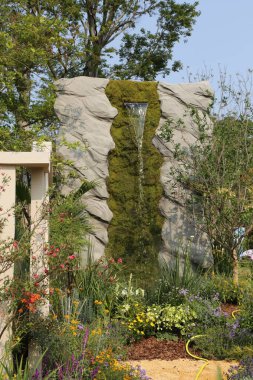 Büyük bir kayanın içinden su akıyordu. Açık bir bahçenin ortasındaydı. Kaya yosunla kaplıdır ve çiçekler ve bitkilerle çevrilidir..