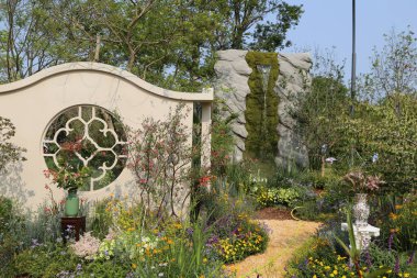 Ağaçlar, çalılar ve çiçeklerle çevrili bir çiçek tarlasının ortasında kemerli bir kapağı ve yuvarlak bir penceresi olan bahçe duvarı, antik kalıntıları hatırlatan bir atmosfer yaratıyor..