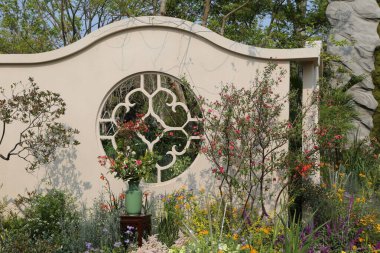 Bahçenin beyaz duvarı kemer şeklinde bir pencere ve çiçeklerle süslenerek üzerinde dairesel bir desen oluşturulur. Arka planda, çeşitli bitkiler ve renkli kır çiçekleriyle çevrili doğal ışık altında ağaçlar yer alıyor.