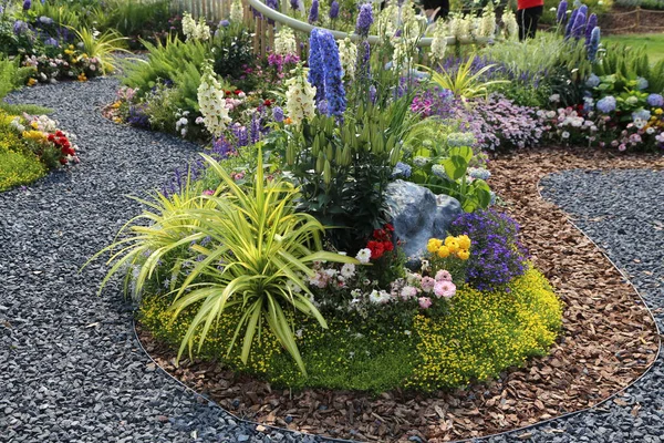 Mavi ve mor sümbüllerle, sarı yapraklarla, kırmızı sardunyalarla, yeşil otlarla ve beyaz zambaklarla çevrili açık bir bahçenin ortasında büyük, yuvarlak bir çiçek yatağı..