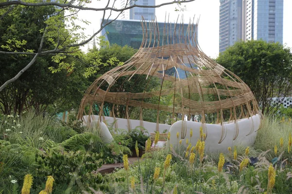 Beyaz duvarları ve yeşil çatısı olan, çeşitli bitki ve çiçeklerle çevrili, yumurta şeklinde bambu bir pavyon. Önünde arkasında yüksek binalar olan bir şehir parkı var..