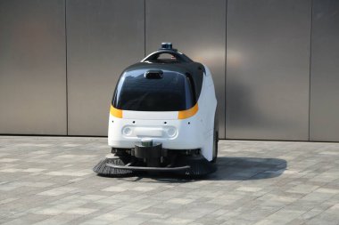 Sarı aksanlı beyaz ve siyah süpürge bir robot açık hava otoparkının zemininde çalışıyor, ön görüş, tüm gövde çekimi, endüstriyel tasarım, temiz arka plan, doğal ışıklandırma
