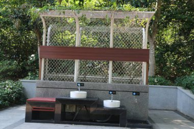 Önünde iki lavabo, arkasında da açık hava olan açık bir tuvalet. Her birimin duvarlarında tel örgü kapılar var. Önlerinde de bir bank var, beklemek için..