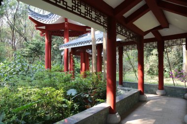 Parkta yeşil bitkilerle çevrili kırmızı sütunları ve saçakları olan Çin tarzı bir pavyon. Toprak beyaz taş levhalarla kaplı..