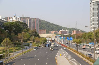 Solunda binalar ve yeşillikler olan bir otoyol, iki yönde de giden arabalar. İki şehri birbirine bağlayan bir üst geçit var..