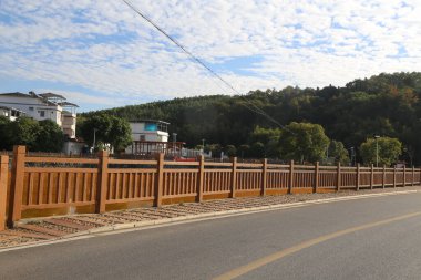 Yolun ahşap korkulukları bireysel el yapımı kereste korkuluklarından oluşuyor, Çin kırsalında bir caddenin her iki tarafında zarif bir sokak çiti oluşturuyorlar ve arka planda yeşil dağlar ve beyaz bulutlar var.