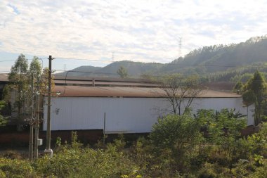 a white warehouse with two floors, an old roof, and a large building next to it on the hillside in a rural area of china. the photo was taken from outside through a car window, showing green vegetation all around.