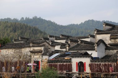 Köyde bir grup sazdan ev, beyaz duvarlar ve siyah fayanslar, ve üzerinde kırmızı bez asılı. Kurutulmuş biberler binanın dışında ve arka planda dağların arkasında ağaçlar görülebiliyor..