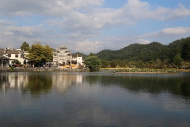 Çin kırsalında her iki tarafında binalar ve ağaçlarla çevrili güzel bir göl. Önünde beyaz duvarları ve siyah karoları olan antik bir bina var. Sakin sulara yansıyor.