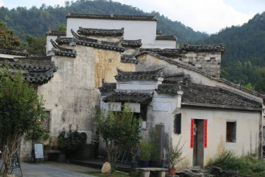 Anhui 'de bir kırsal ev, beyaz duvarları ve gri fayansları olan bir porselen, dağlarla ve yeşil bitkilerle çevrili. Çatı, bir duvarın kenarından görülebilen siyah kiremitlerle kaplıdır..