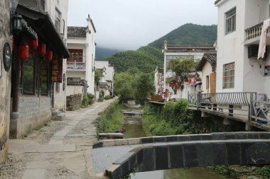 Caddenin her iki tarafında beyaz duvarlar ve siyah fayanslarla antik Anhui kasabasından akan küçük bir dere. Uzakta yeşil dağlarla çevrili eski ahşap bir köprü var..