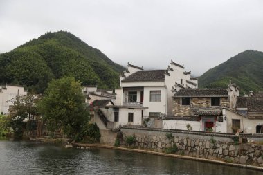 Bir grup eski beyaz Çin binası nehir kıyısında, yeşil dağlarla çevrili, Çin kırsalında bulutlu bir gökyüzü olan ağaçlarla çevrili. Sahne geniş açılı çekimler, her iki tarafta taş duvarlar, basit evler, uzak yerlere uzanan taş yollar içeriyor.