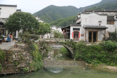 Yolun her iki tarafında beyaz duvarlar ve yeşil fayanslar olan eski bir Çin köyünde nehrin üzerinde küçük bir köprü. İnsanlar ağaçların altında oturmuş, suyun akışını izliyorlar..