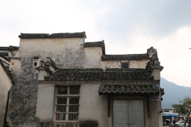 Beyaz duvarları ve siyah kiremitleri olan kırsal bir ev. Çatı zamanla hasar görmüş. Hemen yanında, ön duvarının bir tarafında küçük pencereleri olan eski gri bir bina var. Bu binaların önünde, Çin 'deki dağlara giden bir yol var.