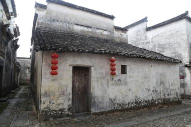 Duvarında kırmızı fenerler asılı beyaz duvarlı, gri fayanslı kırsal bir ev ve önünde antik bir sokak. Duvarlar zaman içinde rüzgar, yağmur, güneş ve elementler yüzünden yıprandı..