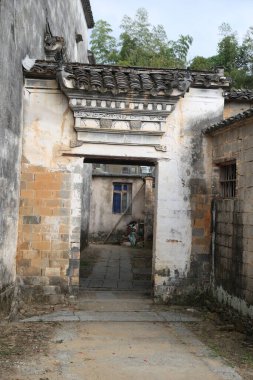 Yıpranmış taş ve tuğla kemer yolu geleneksel Çin köyü girişini oluşturuyor. Döşemeli çatı ve soluk alçı gösteri yaşı. Arka avlu mavi bir kapıya çıkıyor. Olay yerini ağaçlar ve yapraklar çevreledi
