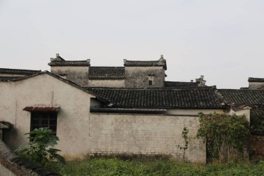 Çin köyü, beyaz duvarlar ve basit mimarisi olan siyah fayanslı evler. Çatı gri kiremitli çatılarla kaplıdır. Yerde yeşil bir bitki örtüsü var ve bir evin arkasındaki eski taş duvarın bir kısmını görebilirsiniz..