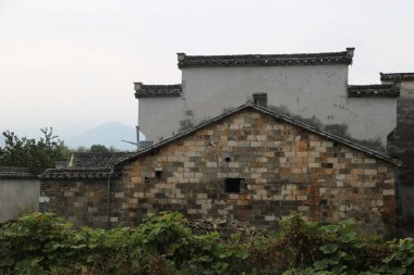 Eski tuğla duvar ve çatısı olan, gri kiremitlerle kaplı kırsal bir ev, diğer iki evin sol tarafında yer alıyor. Altında yeşil bitkiler var..