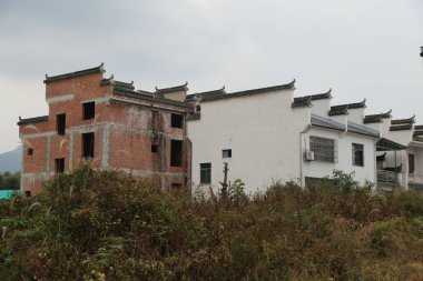 İki binanın solunda beyaz duvarlar bulunan kırsal tuğla ve beton bir ev yapım aşamasında. Diğer binada kırmızı tuğlalar ve siyah fayanslar var. Önünde yeşil bir bitki örtüsü var.