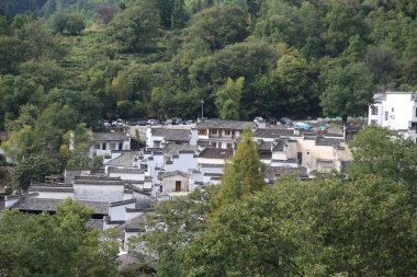 Tarihi bir kırsal köyün yoğun ve üst üste binen çatılarına odaklanan ayrıntılı bir bakış açısı. Klasik Huizhou mimarisi, el değmemiş beyaz cepheleri ve koyu gri fayanslarıyla, yemyeşil yapraklarla güzel bir tezat oluşturarak, bir gli sunar.