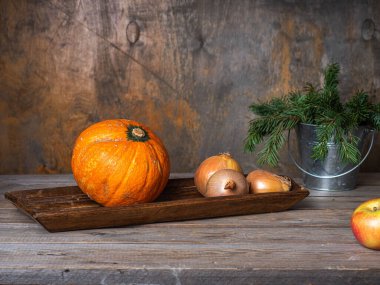 Portakal kabağı ve köknar dalları teneke kovada, ahşap tepside çiğ sebzeler ve basit bir mutfak masasında.