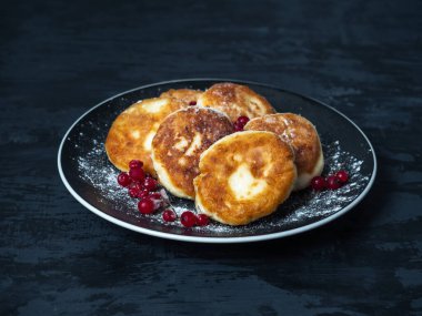 Delicious dessert, syrniki with powdered sugar and red currant berries, serving on a black kerpmic plate on a dark background