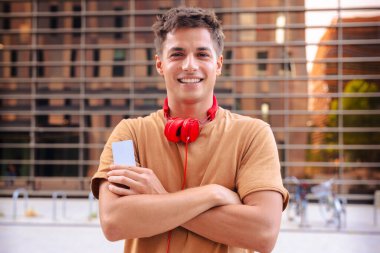 Kollarını kavuşturup kameraya gülümseyen, boynuna kırmızı kulaklık takan şehir arka planında duran, sıradan moda tarzı, kendine güvenen bir genç adam.