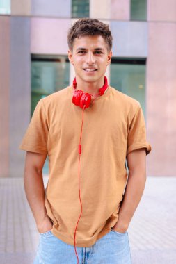 Boynuna kahverengi tişört ve kırmızı kulaklık takmış genç bir adam, gülümseyen bir ifadeyle doğrudan kameraya bakıyor, modern kentsel bir arka planın önünde dikey duruyor.