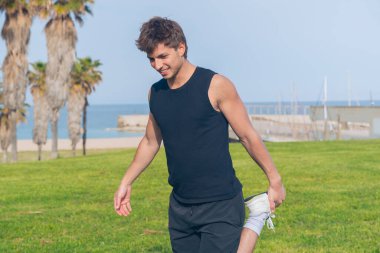 Atletik genç adam, plaj parkında egzersiz yapmadan önce yeşil çimlerin üzerinde bacağını esnetiyor, kasları egzersiz için hazırlıyor, sağlıklı yaşam tarzı, fitness eğitimi, aktif yaşam, motivasyon, dayanıklılık, denge.