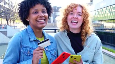 İki mutlu çok ırklı kadın ellerinde akıllı telefonlar ve kredi kartı, çevrimiçi alışveriş konsepti, dijital ödeme, arkadaşlık, eğlence, modern yaşam tarzı, sosyal bağlantı, moda insanlar