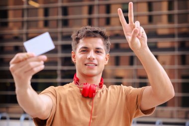 Gülümseyen genç adam kameraya doğru boş bir beyaz kart tutarken dışarıda kırmızı kulaklıklı barış işareti gösteriyor, günlük tarz, kentsel arka plan, model, reklam, kopyalama alanı konsepti