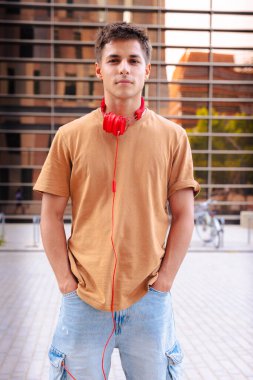 Boynuna kahverengi tişört ve kırmızı kulaklık takmış genç bir adam, kameraya ciddi bir ifadeyle bakıyor, modern kentsel bir arka planın önünde dikey duruyor.