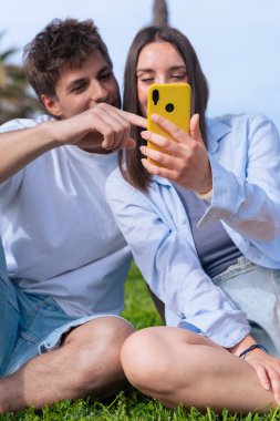 Güneşli bir parkta eğlenen mutlu genç çift, erkeğin kadının elinde tuttuğu akıllı telefon ekranını işaret ederken gülümsüyor. Palmiye ağaçları ve açık hava manzarası rahatlık yaratır. 