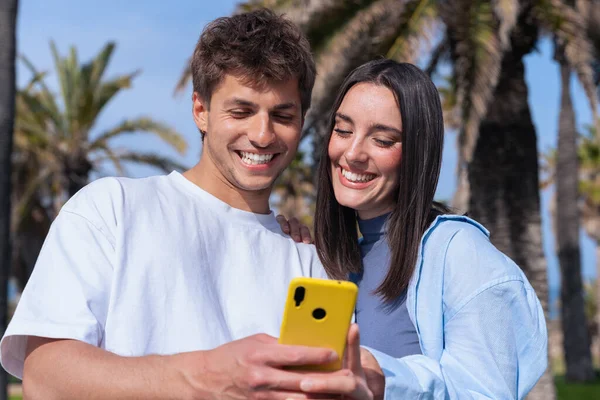 Güneşli parkta birlikte akıllı telefona bakan mutlu genç çift. Palmiye ağaçları, aşk, iletişim, teknoloji, ilişki, sosyal medya, mutluluk, tatil, açık hava yaşam tarzı..