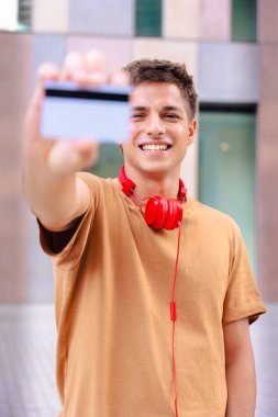 Boynunda kırmızı kulaklıklarla gülümseyen genç bir adamın portresi ön planda bulanık bir şehir geçmişiyle pozitif ve modern bir tutum sergiliyor. Dikey