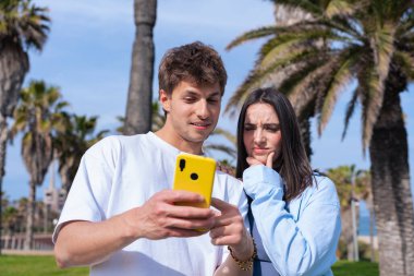 Sunny Park 'ta tuhaf ifadeli akıllı telefona bakan genç bir çift, teknoloji, iletişim, sürpriz, dijital yaşam tarzı, duygu, keşif ve sosyal medya etkisi bugün