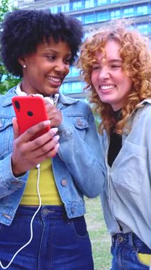 İki neşeli, çok ırklı genç kadın şehrin dışında birbirlerine yakın duruyorlar, akıllı telefon ekranına bakıp gülüyorlar, rahatlamış dikey portre ile eğlenceli sosyal medya içeriklerini paylaşıyorlar.