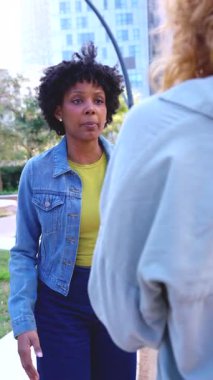 Şehir parkında yüz yüze duran iki genç kadın tartışıyor, biri ciddi bakışlarla hayal kırıklığını ifade ediyor, günlük kıyafetler giyiyor, anlaşmazlık ve gerilimi temsil ediyor..