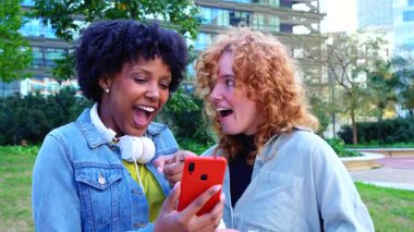 İki mutlu bayan arkadaş, akıllı telefonlar kullanarak, sosyal medya uygulamalarını kontrol ederek, şehir parkında oturarak, gülümseyerek, arkadaşlıkla, modern teknolojiyle, iletişimle, yaşam tarzıyla, 