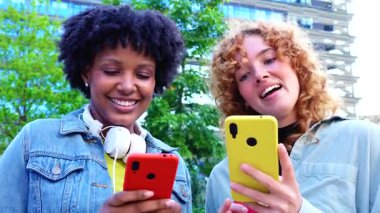 İki mutlu bayan arkadaş, akıllı telefonlar kullanarak, sosyal medya uygulamalarını kontrol ederek, şehir parkında oturarak, gülümseyerek, arkadaşlıkla, modern teknolojiyle, iletişimle, yaşam tarzıyla, 