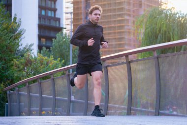 Şehir köprüsünde koşan atletik bir adam siyah spor giyimli, odaklanmış ve kararlı bir ifade takınıyor. Arka planda şehir binaları ve ağaçlar var. Açık hava fitness, ve sağlıklı yaşam tarzı motivasyon konsepti.
