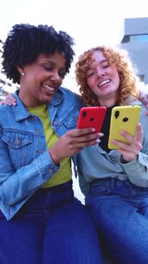 İki genç çok ırklı kadın, dışarıda dikey bir şekilde oturuyorlar, gülümsüyorlar ve vakit geçiriyorlar. Sosyal medyaya akıllı telefondan bakıyorlar, eğlenceli anları paylaşıyorlar ve gündüz vakti arkadaşlıklarını rahatlatıyorlar..